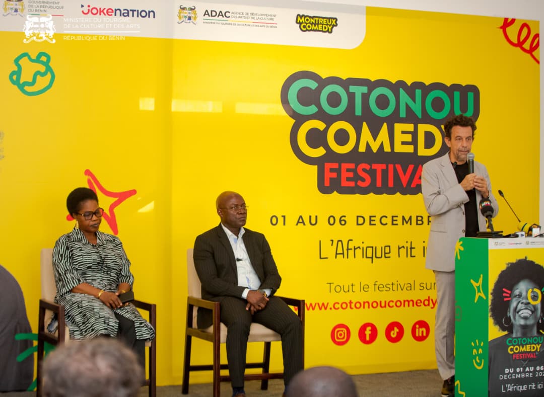Trois personnes lors d’une conférence avec un homme debout au micro et deux assis devant un mur jaune avec le logo du festival Cotonou Comedy