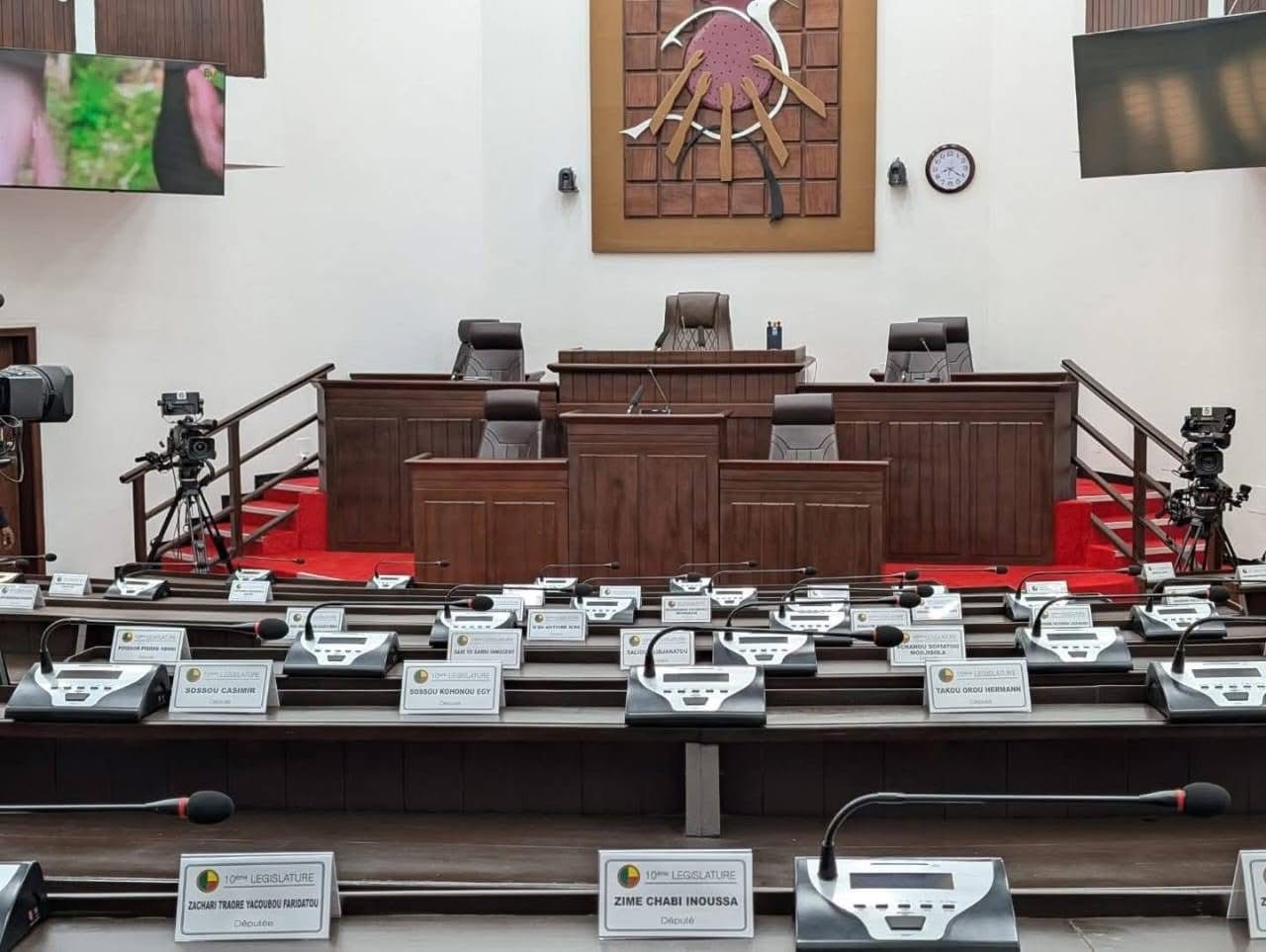 Salle d'assemblée parlementaire avec sièges équipés de micros et étiquettes nominatives vides devant un pupitre central vide