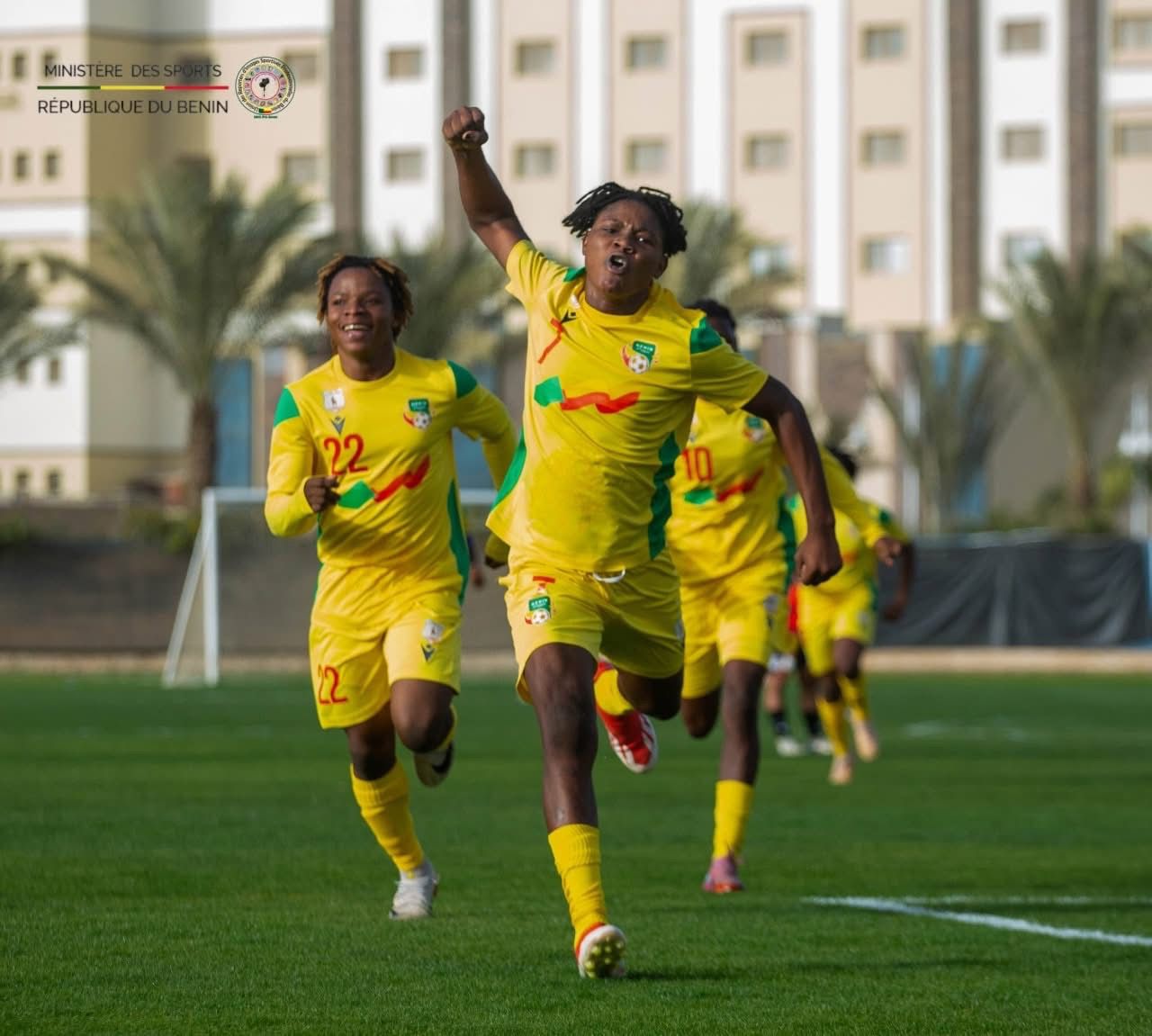 Des joueuses de football en maillots jaunes courent et célèbrent un but sur un terrain herbeux avec des bâtiments en arrière-plan