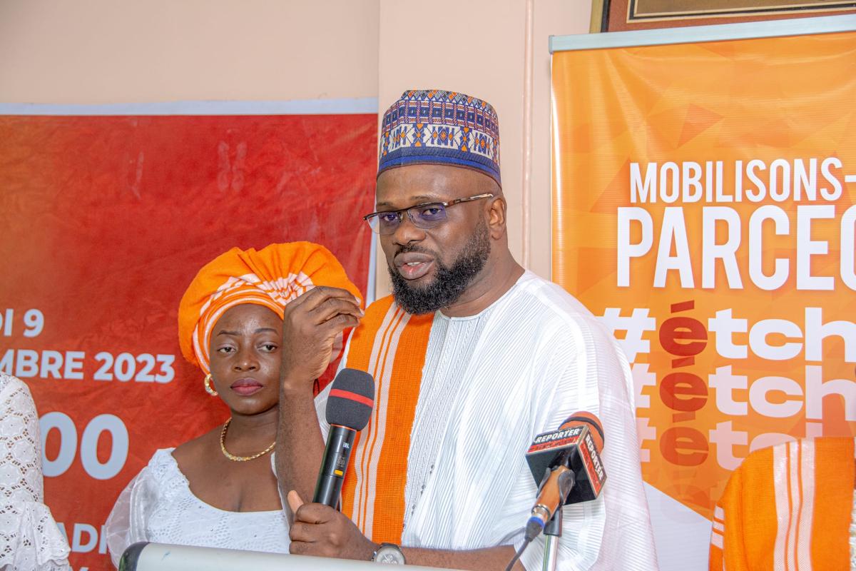 Un homme en tenue traditionnelle blanche et orange parle au micro devant un panneau orange tandis qu'une femme en tenue blanche et coiffe orange l'écoute.