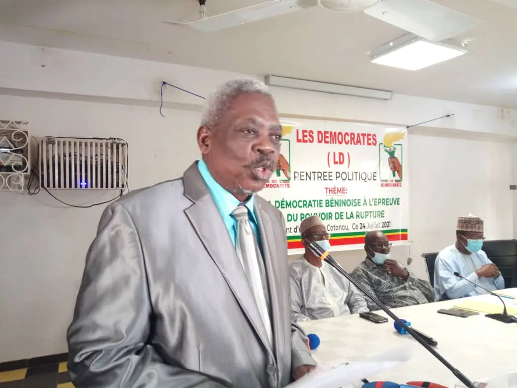 Un homme en costume gris parle au microphone devant trois hommes assis, masque facial sur certains, avec une banderole politique en arrière-plan