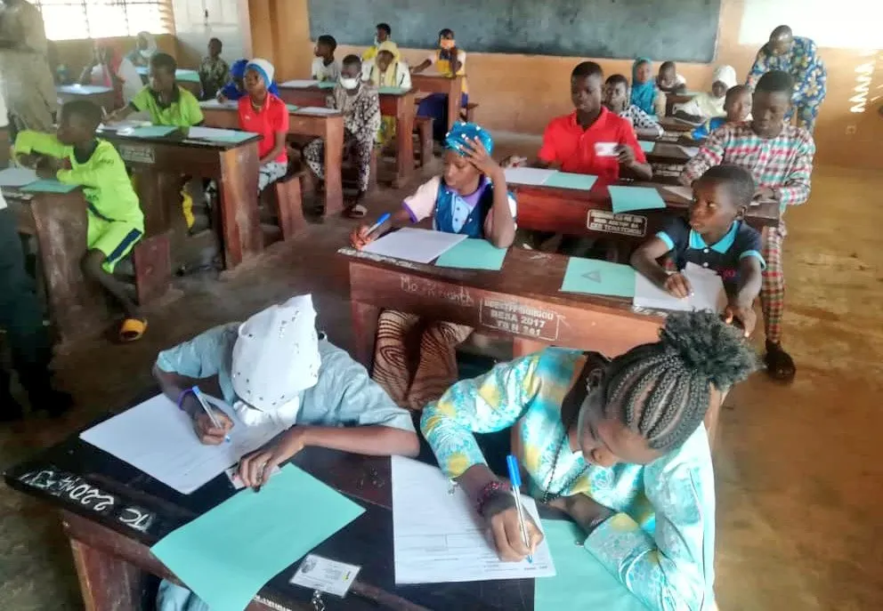 Des élèves assis à des bureaux en bois écrivent sur des feuilles avec des stylos dans une salle de classe éclairée par une fenêtre