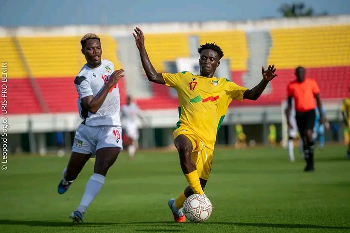 Deux joueurs de football en action : l’un en maillot jaune contrôle le ballon devant un adversaire en blanc dans un stade presque vide