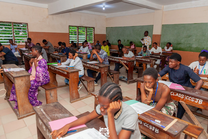 Classe avec plusieurs élèves assis aux bureaux en bois concentrés sur des feuilles d’examen dans une salle d’école avec tableau vert