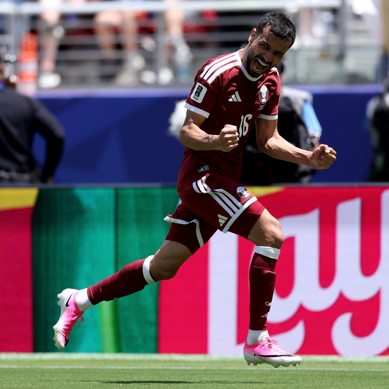 Joueur en maillot rouge foncé célèbre un but en serrant les poings sur un terrain de football en plein jour