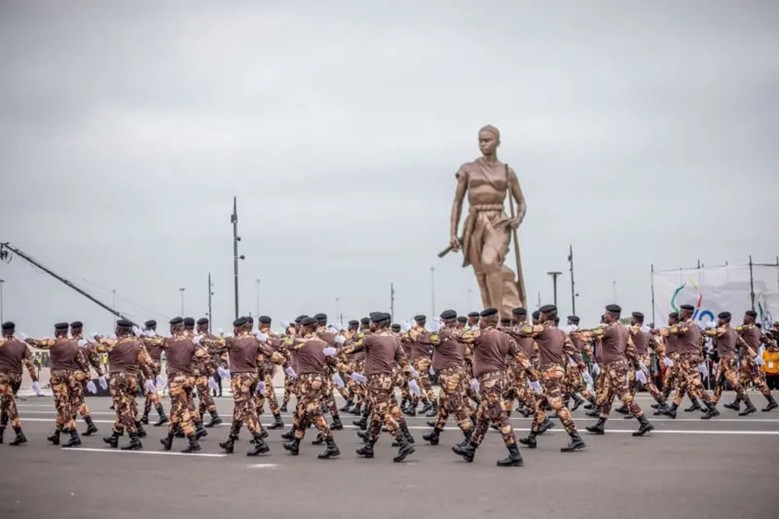 Des militaires en uniforme marron et camouflage marchent en formation devant une grande statue humaine en bronze sur une place ouverte