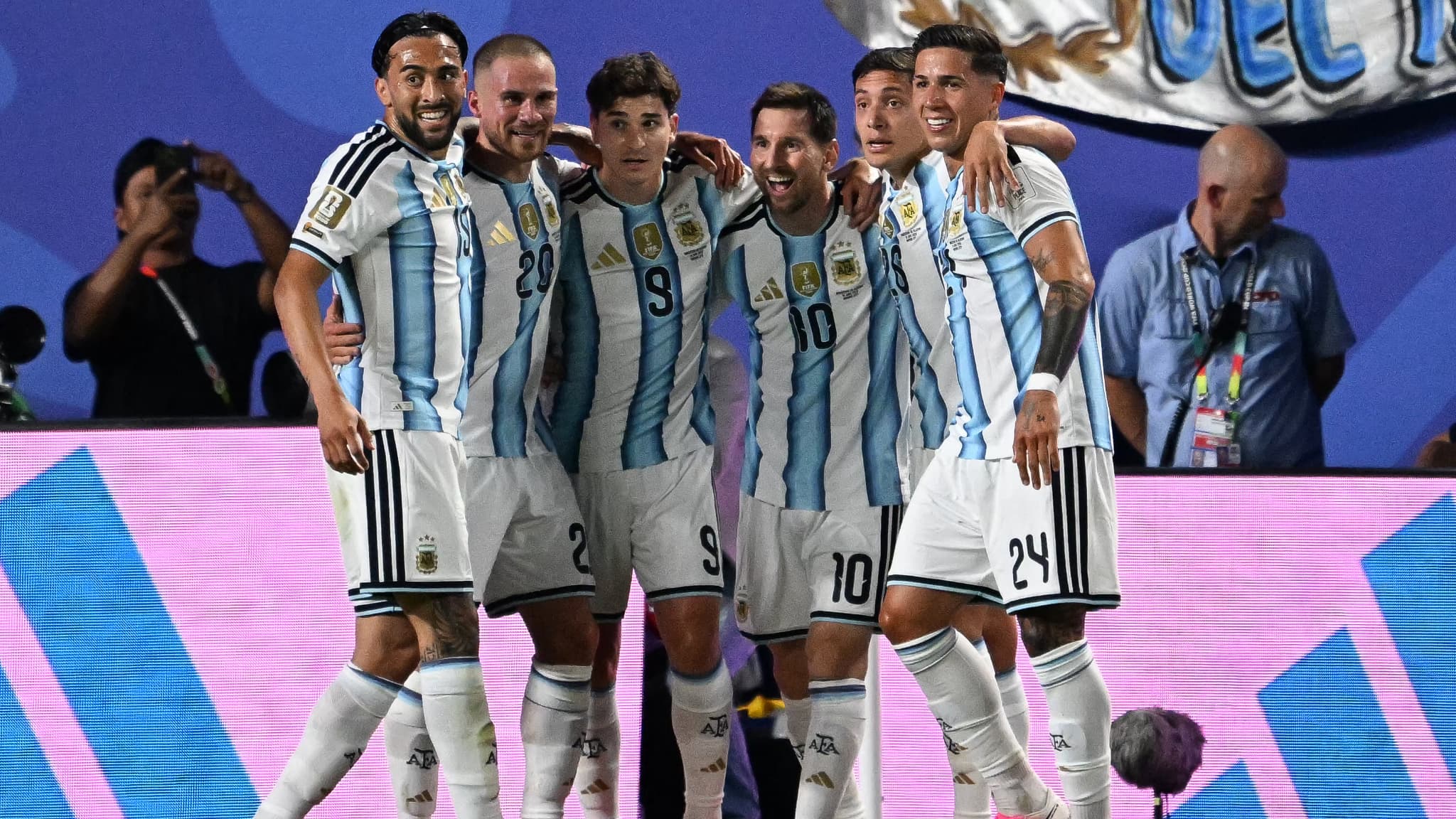 Cinq joueurs de football en maillots rayés bleu et blanc se tiennent bras dessus bras dessous sur un terrain de jeu devant un écran lumineux.