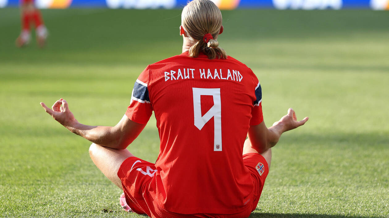 Un joueur en maillot rouge assis en tailleur sur un terrain de football, vu de dos avec les bras ouverts.