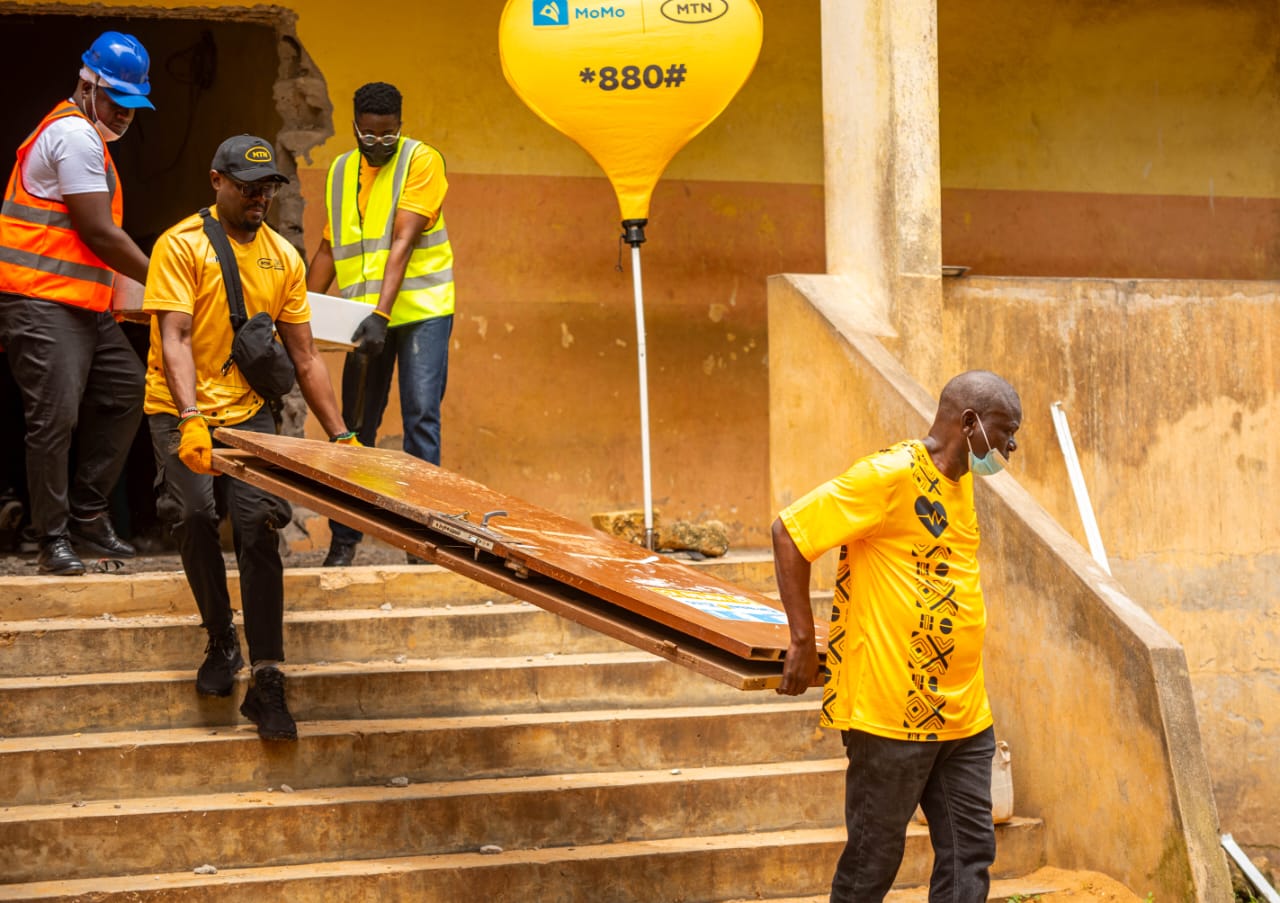 Quatre hommes portant des gilets de sécurité transportent une grande porte en bois dans un escalier extérieur d’un bâtiment jaune
