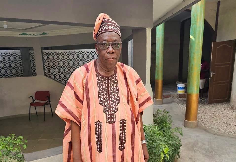 Homme âgé en tenue traditionnelle colorée avec un chapeau assorti debout dans une cour avec végétation et colonnes peintes