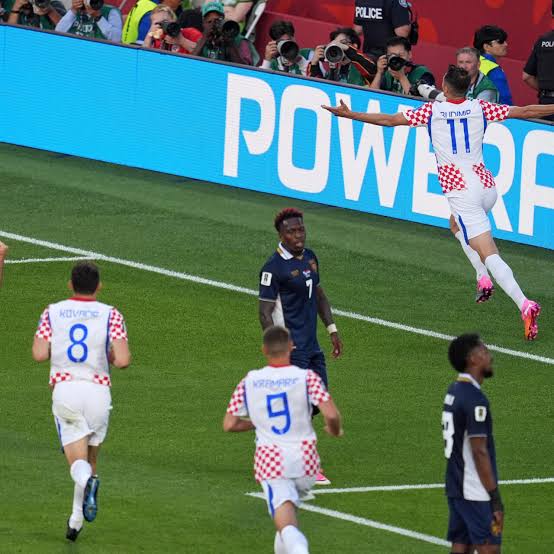 Joueur en maillot blanc à damiers rouges s'élance en l'air pour célébrer un but devant des adversaires en bleu marine et des photographes au bord du terrain