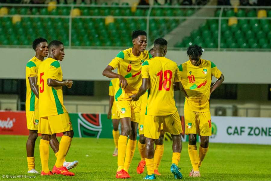 Cinq joueurs en maillot jaune discutent et s’apprêtent à jouer sur un terrain de football avec des gradins verts en arrière-plan