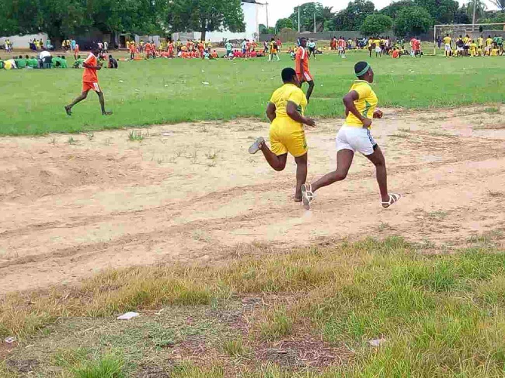 Deux jeunes en tenue sportive jaune courent sur une piste en terre avec d autres compétiteurs et spectateurs en arrière-plan