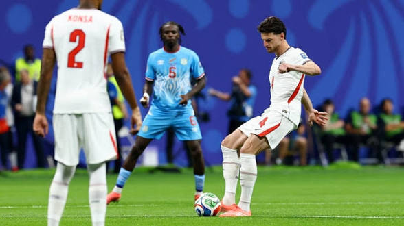 Un joueur en blanc contrôle le ballon face à deux adversaires en maillot bleu sur un terrain de football en intérieur