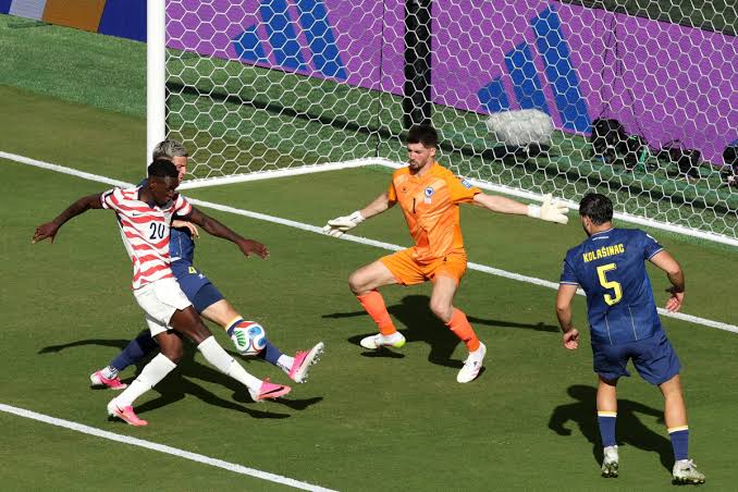 Un joueur en maillot rayé blanc et rouge frappe le ballon devant le gardien en orange et plusieurs défenseurs bleu marine près du but
